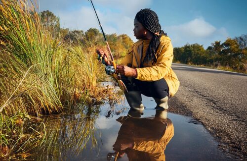 a woman crouching at the edge of a puddle with a fishing pole in her hands