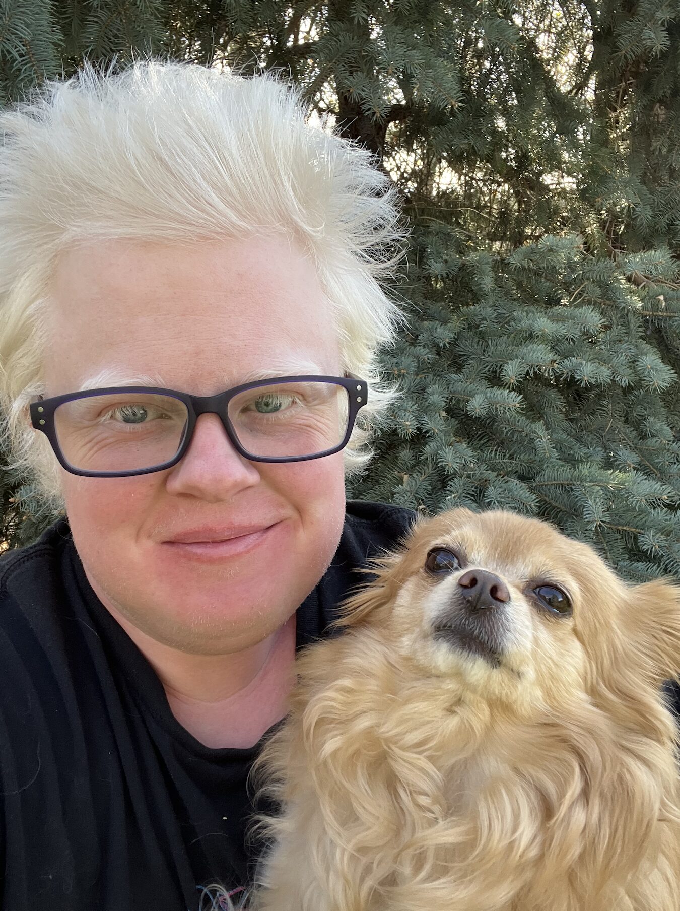 A person with short, platinum blonde hair and black glasses smiles while holding a small, tan long-haired dog in front of green pine trees outdoors.