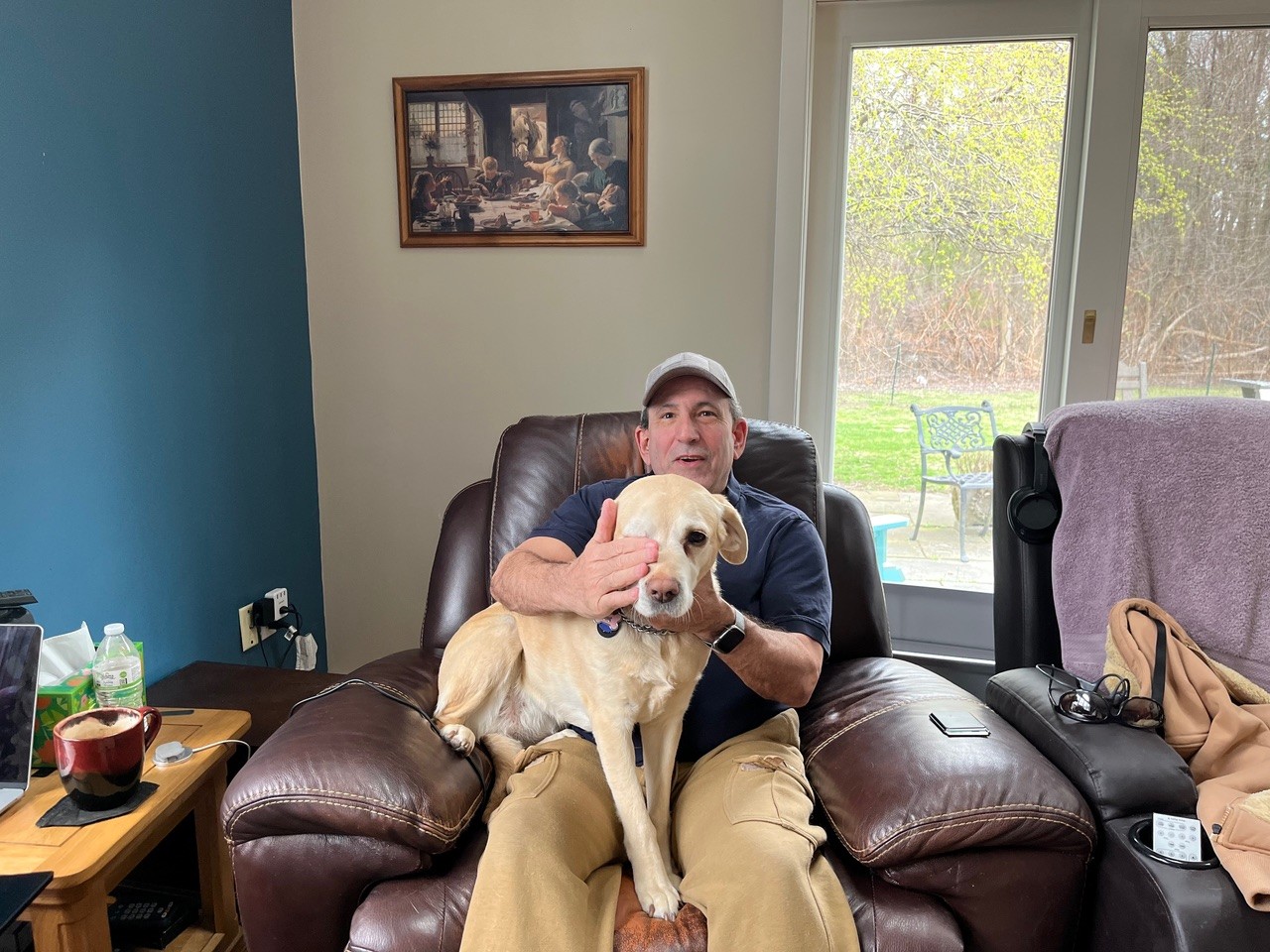 A man sits on a brown recliner in a living room, smiling and covering the eyes of a yellow Labrador dog sitting on his lap. A window shows trees outside, and a framed picture hangs on the wall behind them.