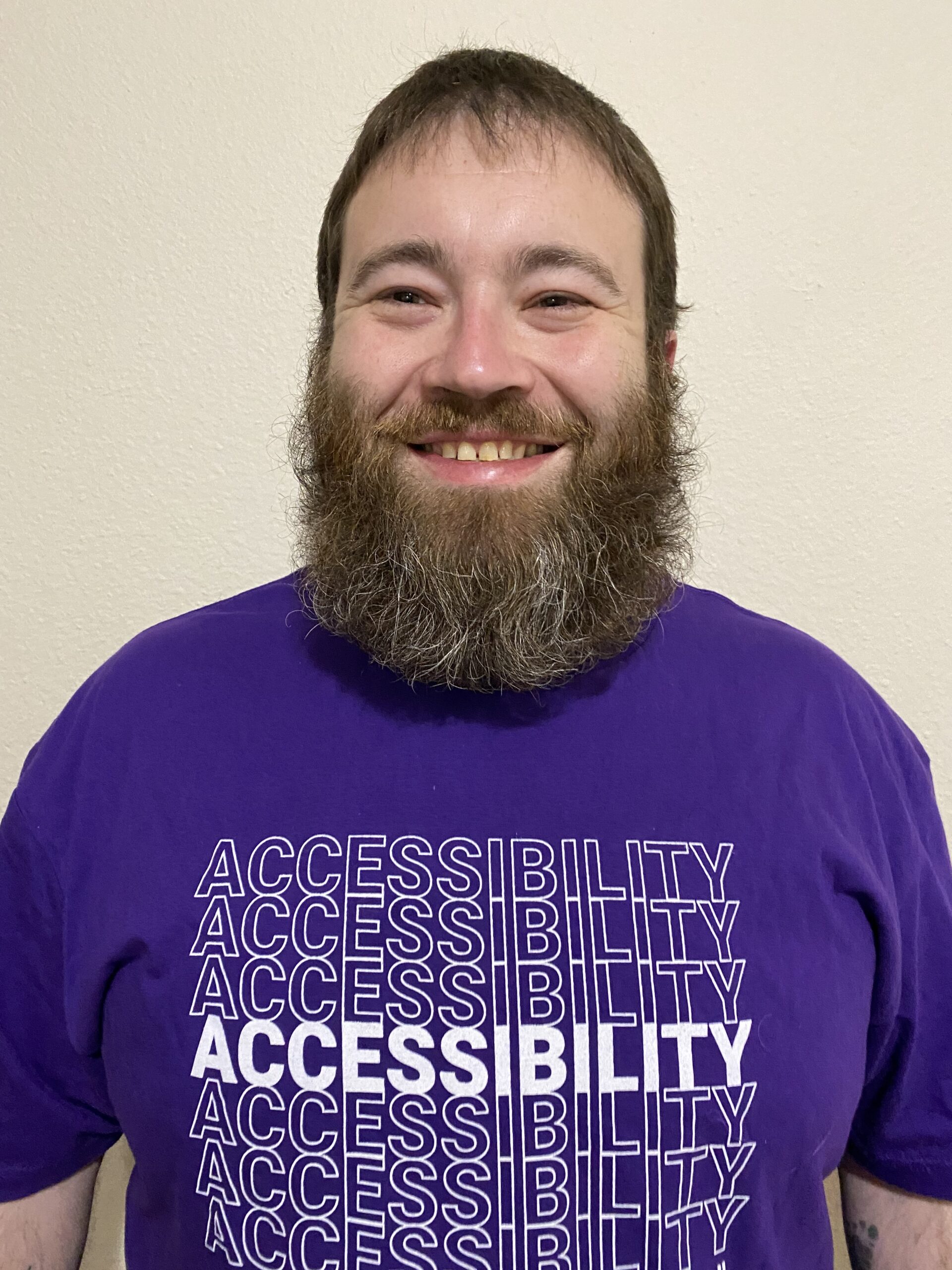 A smiling person with a full beard wears a purple shirt that repeats the word "ACCESSIBILITY" in white text, with "ACCESS" in bold. The background is a plain, light-colored wall.