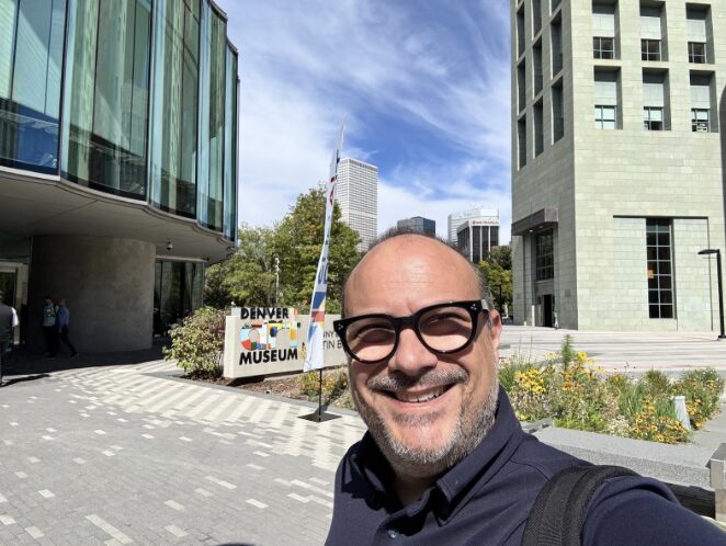 A smiling man with glasses and a backpack takes a selfie outside the Denver Museum. Modern buildings, greenery, and a blue sky with clouds are visible in the background. The museum sign appears behind him, along a paved walkway.