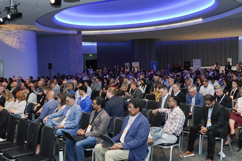 A large, diverse audience sits in rows of chairs at a conference in a modern, spacious room with blue lighting. Many attendees are attentively facing forward, some with laptops or notepads, while a few empty seats are visible near the front.