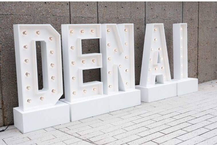 Large white marquee letters spelling “DENIAL” stand on a white tiled surface in front of a gray concrete wall. Each letter is outlined with small round light bulbs, some lit and some unlit, giving a bold, illuminated effect.