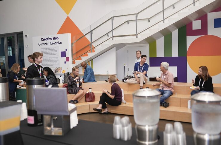 A group of people sit and chat on wooden steps in a modern, colorful room labeled “Creative Hub.” Some hold drinks or food. A counter with pitchers, cups, and beverages is in the foreground, and geometric wall art decorates the background near a staircase.