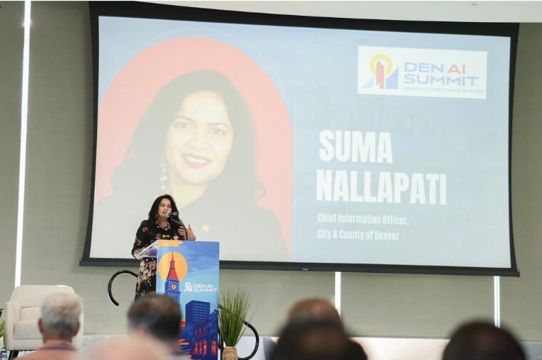 A woman stands at a podium with a microphone, speaking at the DEN AI Summit. Behind her, a large screen displays “Suma Nallapati, Chief Information Officer, City & County of Denver” alongside her photo and the event logo. Audience members are visible in the foreground.