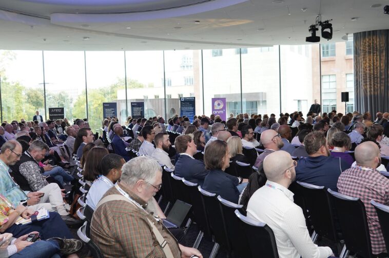 A large audience sits in rows at a conference or seminar in a bright room with floor-to-ceiling windows. Most attendees face forward, listening attentively, while some use electronic devices. Banners and signs are visible in the background.