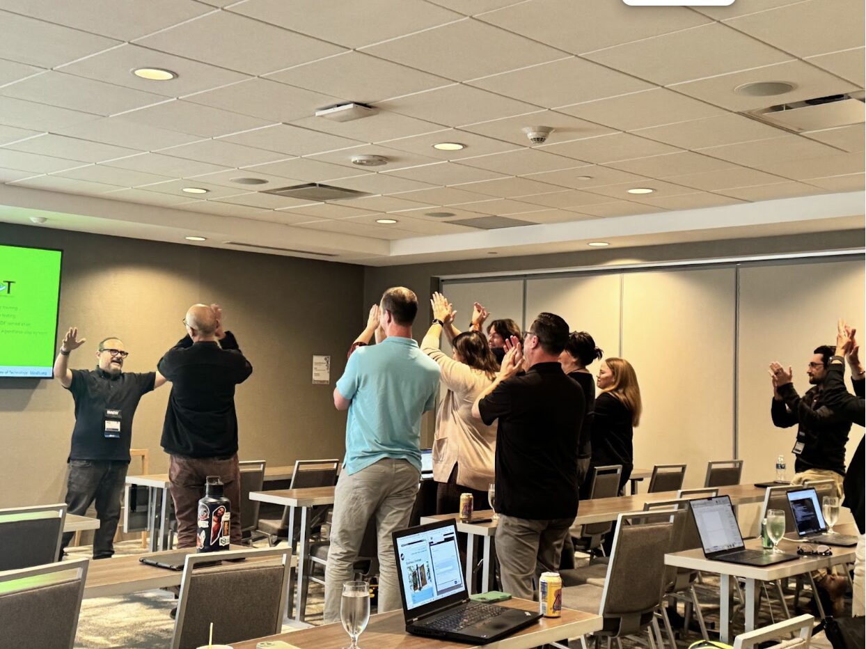 A group of people stand in a conference room, facing a presenter near the front. Everyone has their arms raised in the air, participating in an interactive activity. Laptops and drinks are on the tables, and a presentation slide is visible on the wall-mounted screen to the left. The room has neutral tones and overhead ceiling lights, creating a professional training or workshop environment.