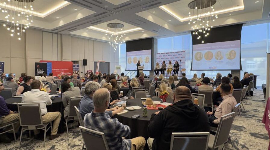 Large metallic balloon letters in blue and red spelling “NAGW” are displayed on a countertop against a wall. Abstract framed artwork hangs above, featuring circular tree-ring-style designs in blue tones. The setup appears to be part of a conference or event decor.
