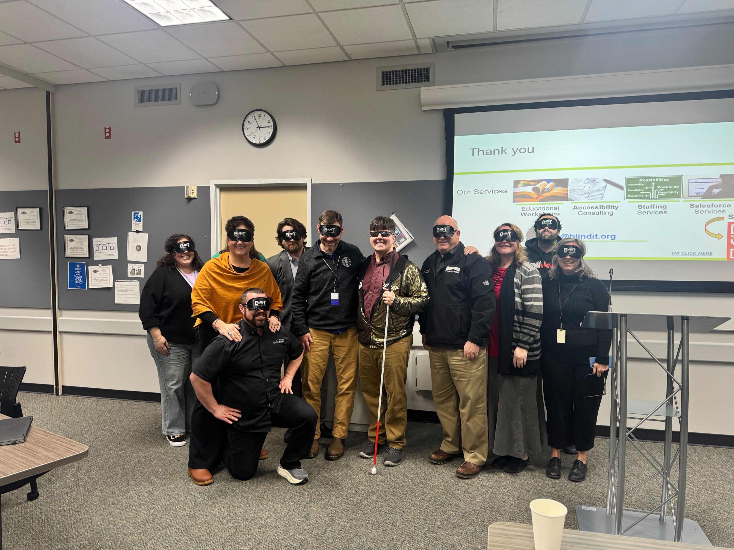 A group of about twelve adults stand together in a classroom or conference room, smiling for a group photo. Most are wearing black blindfolds with white text, and one person in the center holds a white cane. A presentation screen behind them reads “Thank you” and lists services including educational workshops, accessibility consulting, staffing services, and Salesforce services. A podium stands to the right of the group.