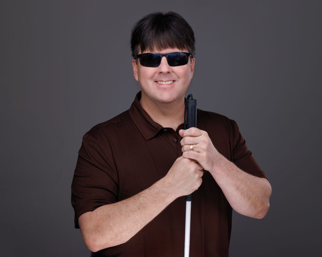 Studio portrait of BIT founder Mike Hess smiling in dark glasses, hands resting on his white cane.
