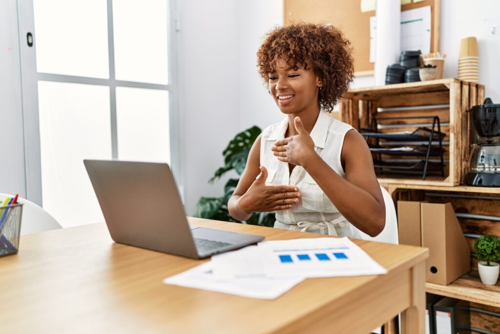 A smiling woman signs to someone on a video call from her laptop in a bright office.