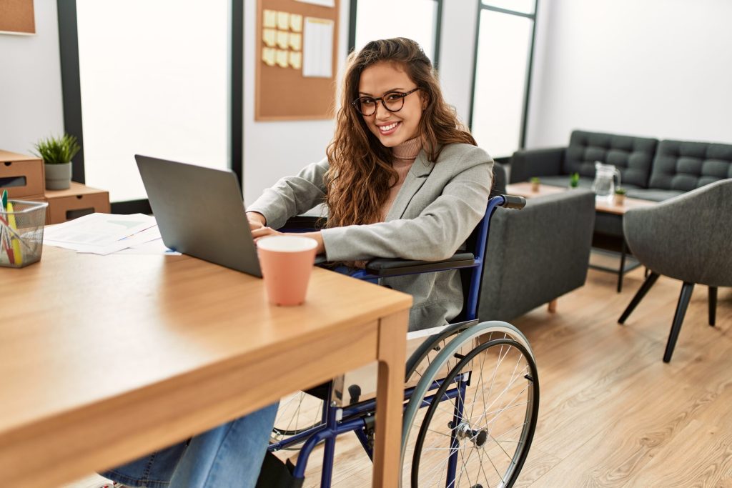 A woman using a wheelchair smiles while working on a laptop at a desk in a bright office.