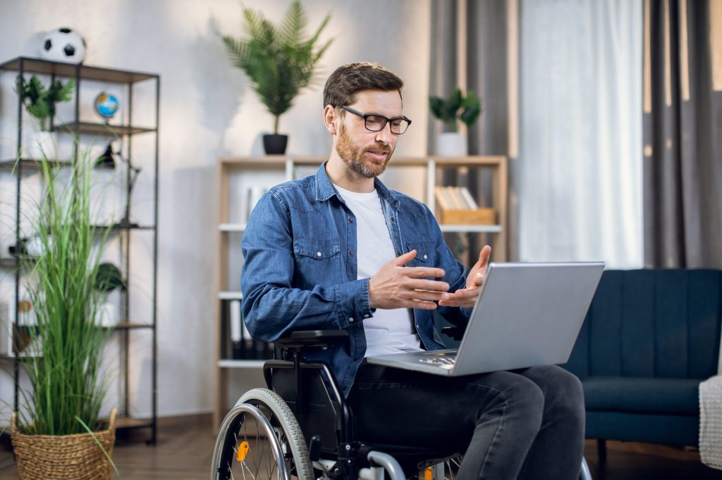 A man using a wheelchair gestures while talking on a video call at his laptop.