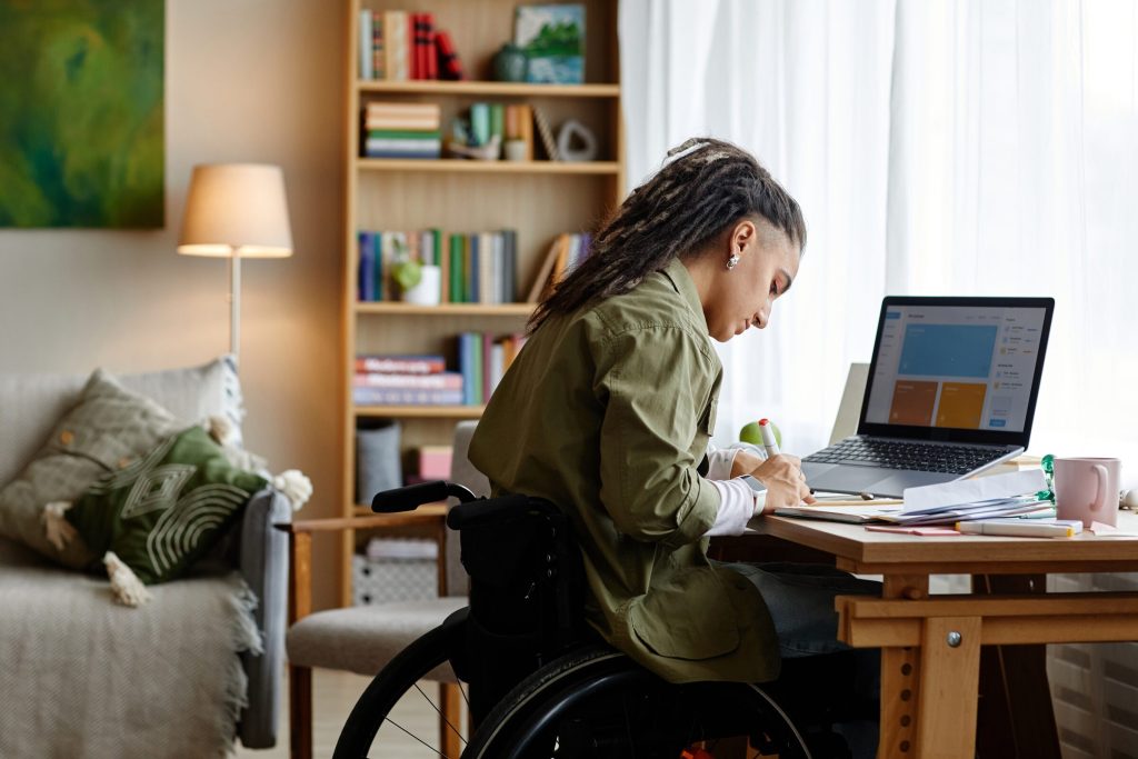 A woman using a wheelchair writes notes at her desk beside an open laptop.