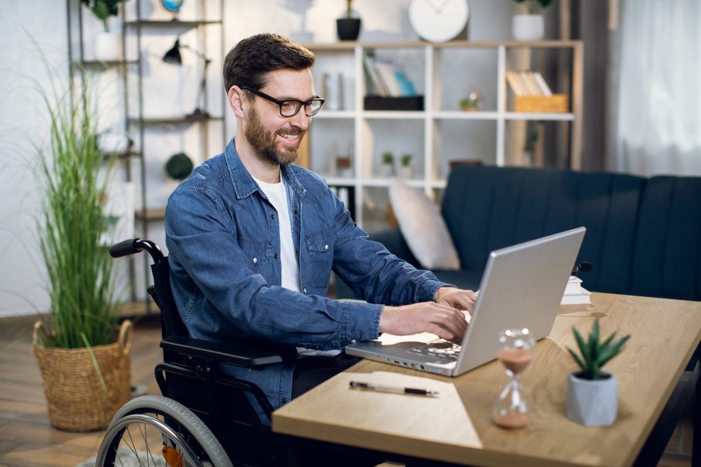 A man using a wheelchair types on a laptop at a wooden desk in a home office.