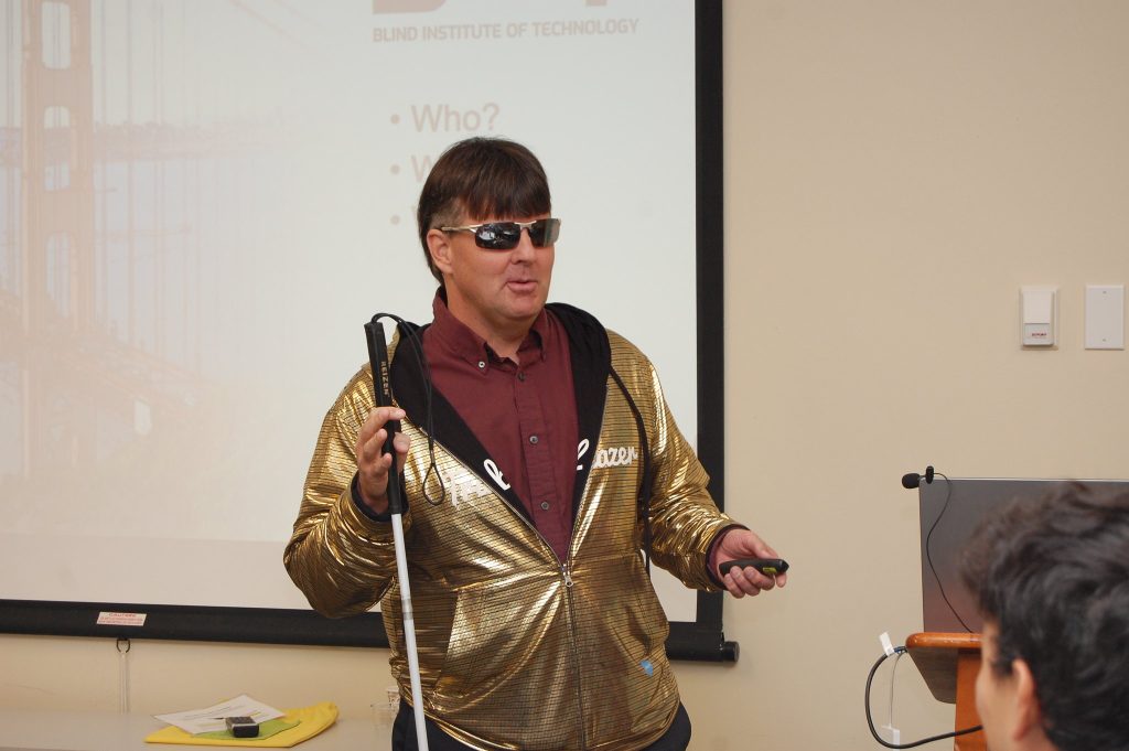 Mike Hess presents in a gold jacket, white cane in one hand and a remote in the other, in front of a Blind Institute of Technology slide.