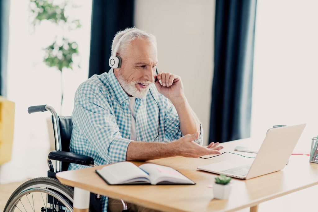 An older man using a wheelchair talks on a headset at his desk, with a laptop and notebook open in front of him.