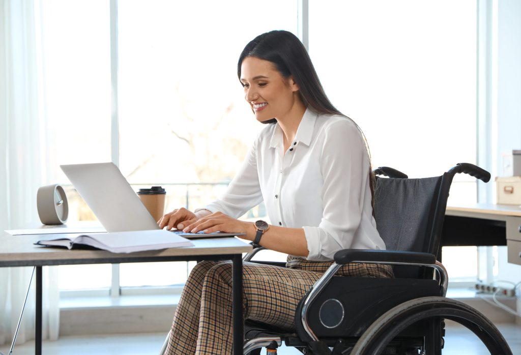 A woman using a wheelchair works on a laptop at a sunlit desk beside a window.